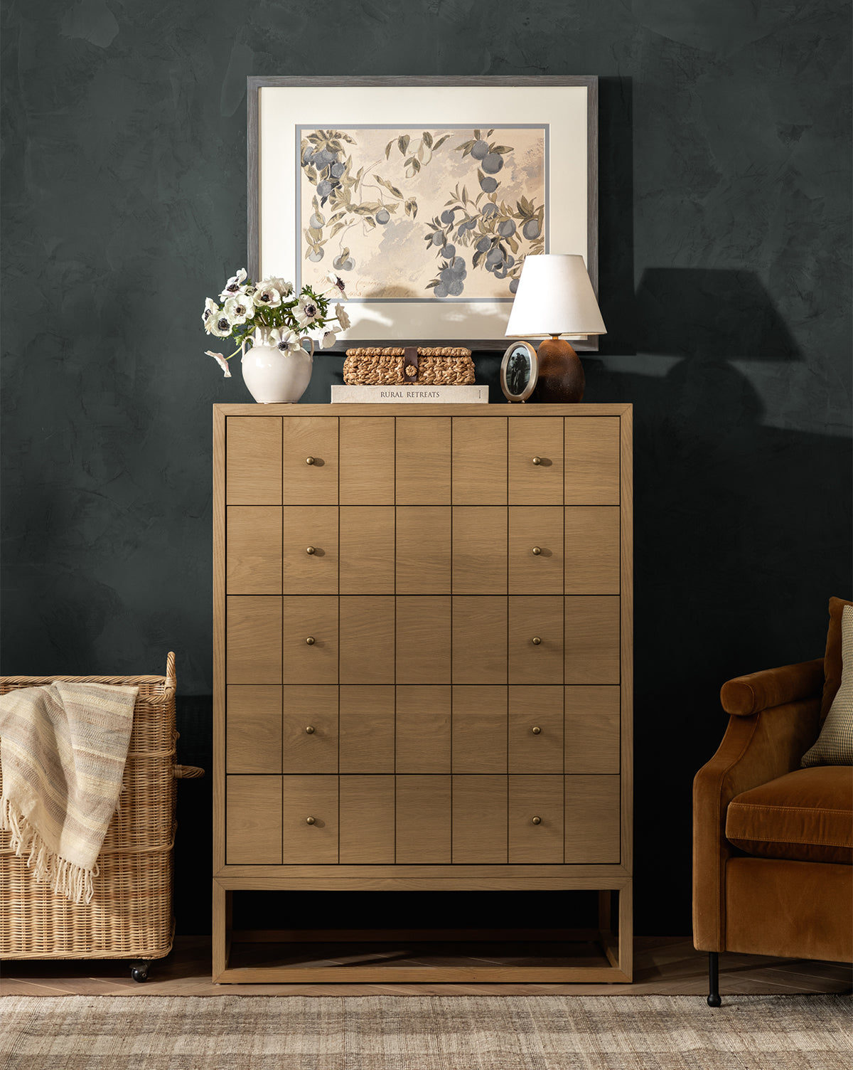 A Huxton Tall Dresser in light wood with a grid pattern stands by a dark wall, topped with floral art, a white lamp, flowers in a vase, and decor. A woven basket and brown chair sit nearby, by McGee & Co.