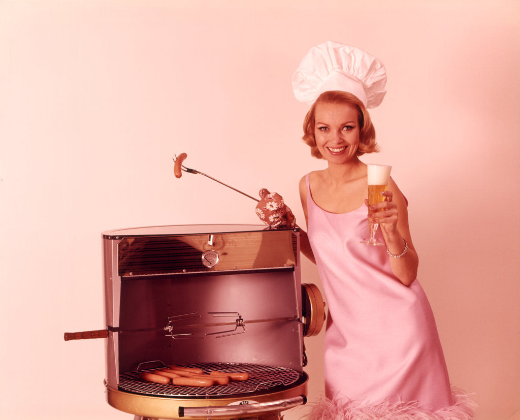 1960s Woman Wearing Pink Party Dress And Chef Hat Grilling Hot Dogs Drinking Beer