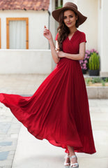 A woman in a flowing red chiffon empire waist wedding dress with cap sleeves, a V-neckline, and a maxi length that reaches the calf. She is wearing a wide brim hat and her dress is flowing in the wind.