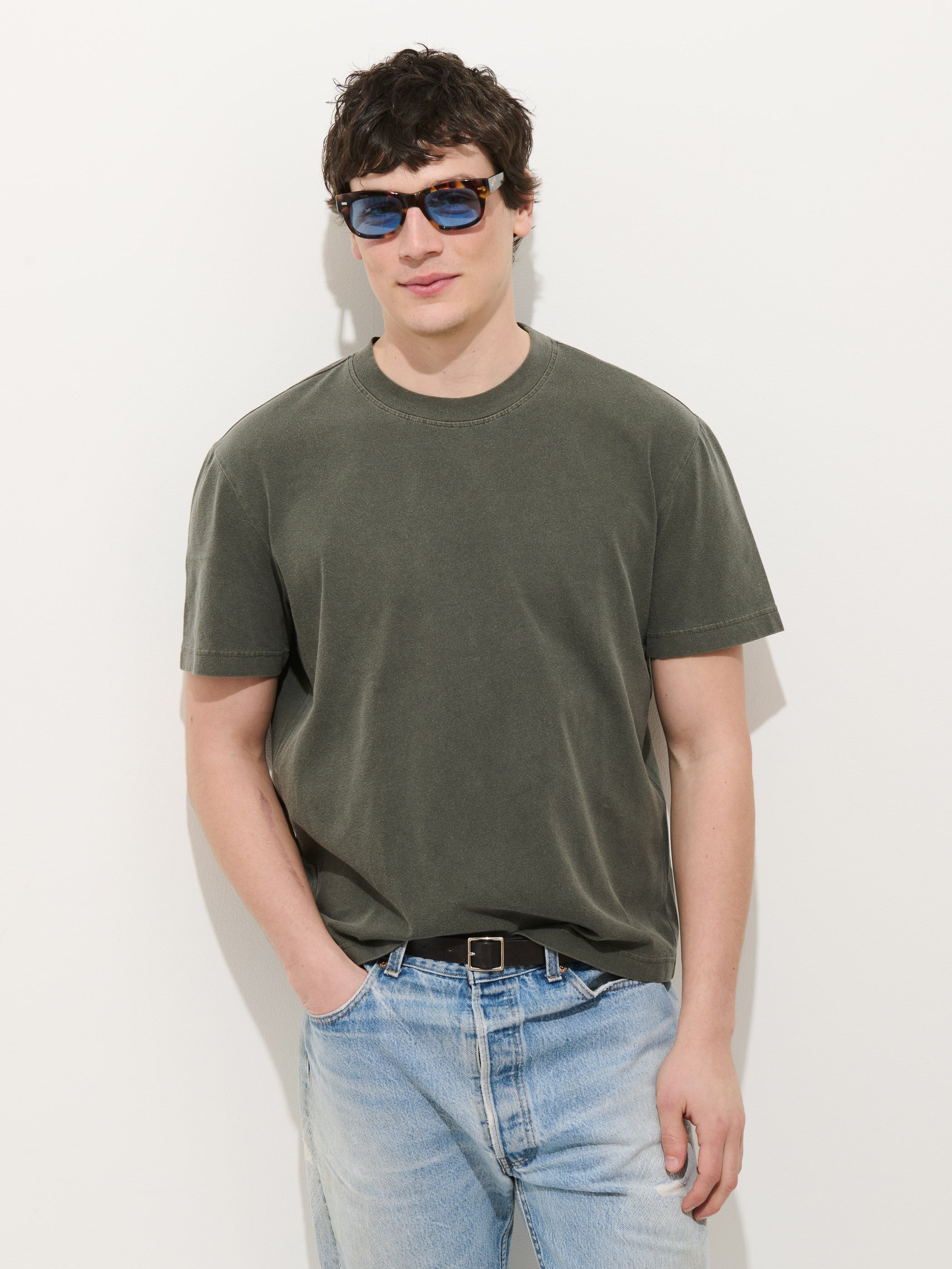 A young man wears blue sunglasses, light blue jeans, and the Crosby Tee in Midweight Jersey—an olive green, 100% cotton garment-dyed shirt—standing against a white background with one hand in his pocket.