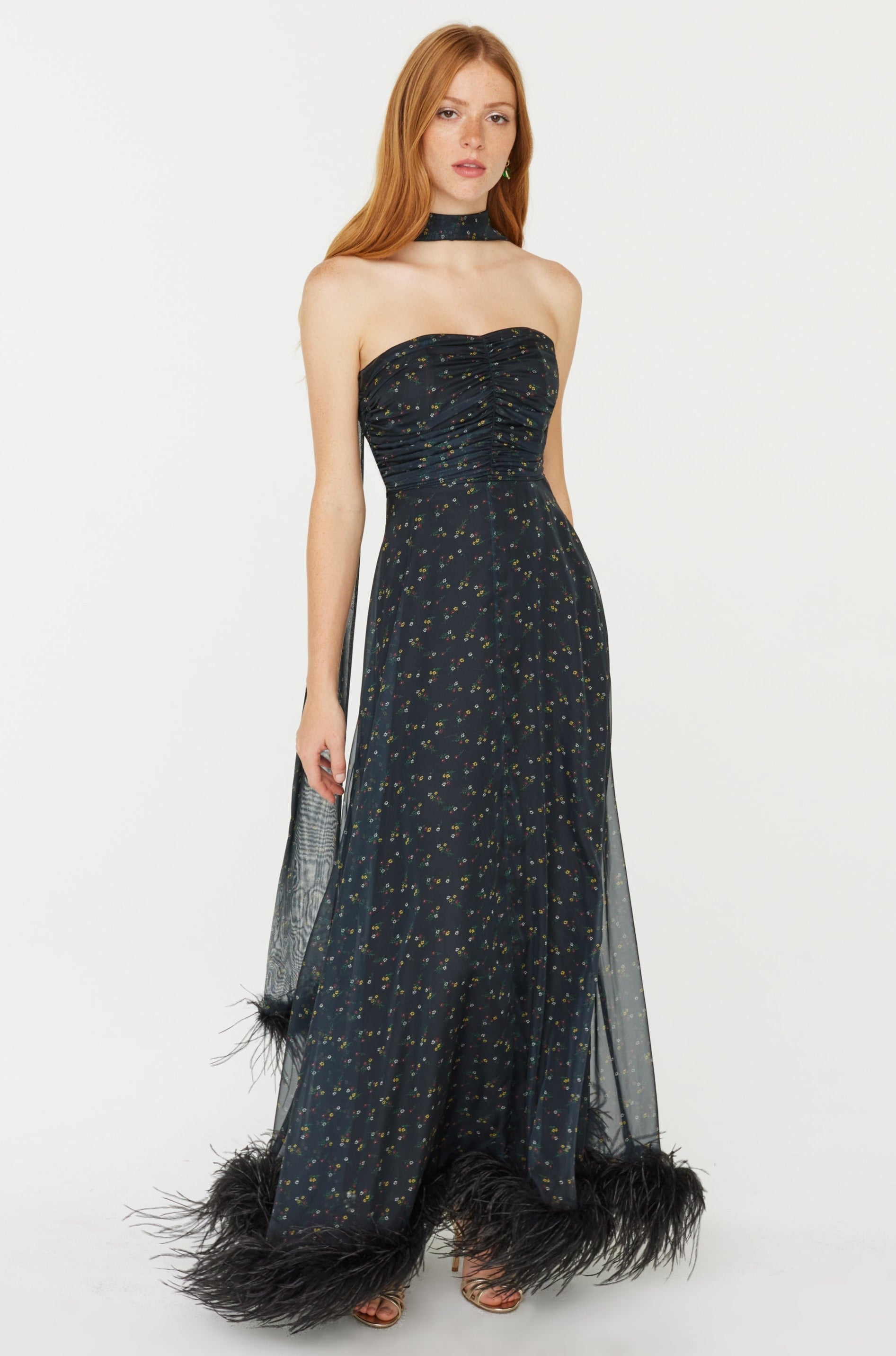 A model wears a strapless corseted chiffon long gown with a matching neck scarf. The dress has black feathers at the hem. The print is black, red, and green small flowers on a black base.