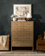 A Huxton Tall Dresser in light wood with a grid pattern stands by a dark wall, topped with floral art, a white lamp, flowers in a vase, and decor. A woven basket and brown chair sit nearby, by McGee & Co.