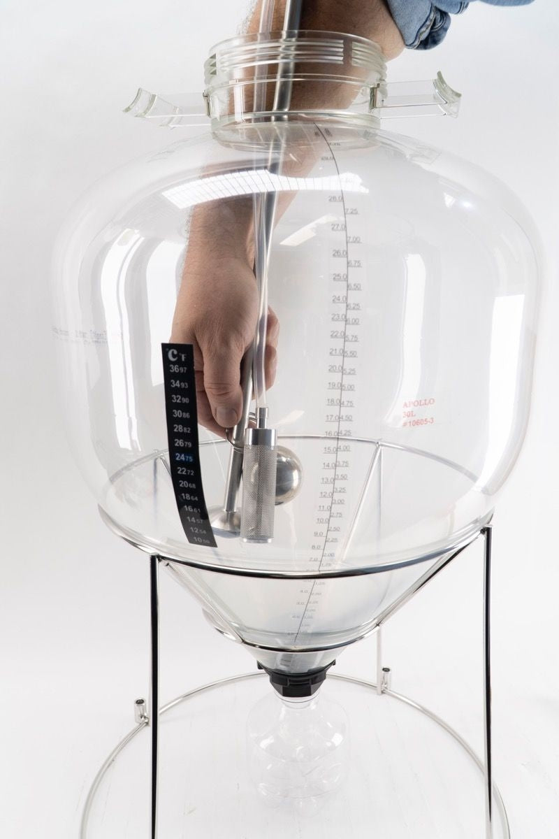 En hånd stikker et termometer og et hydrometer ned i en stor, gjennomsiktig Apollo 30L Unitank - Keg King-fermenter med volumangivelser, støttet av et metallstativ.