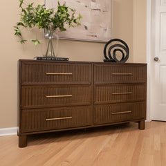 Mid-century modern six-drawer wooden dresser with fluted drawer fronts and brass handles, styled with a glass vase of greenery and abstract black sculpture on hardwood floor against a beige wall