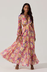The dress features a colorful, watercolor-like floral pattern in shades of pink, purple, yellow, and beige. It has a deep V-neckline, long semi-sheer sleeves with elastic cuffs, and a cutout at the waist that connects to a high-waisted, flowing maxi skirt with pleats. The fabric appears lightweight and airy, creating a soft, movement-filled silhouette. The model is also wearing gold earrings and strappy heels.