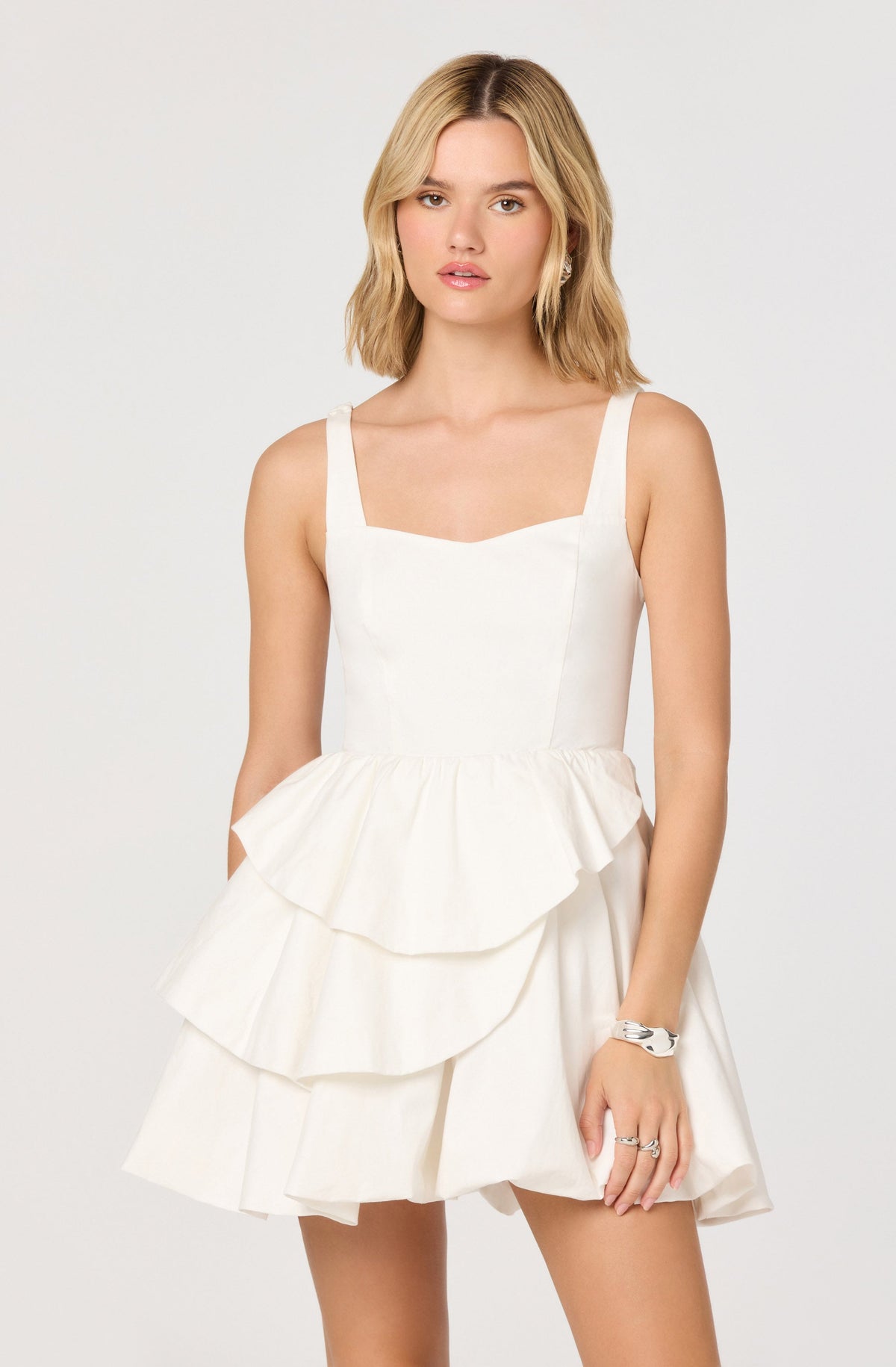 A young woman is wearing a sleeveless, white dress with wide shoulder straps. The dress has a fitted bodice and features layered ruffles on the skirt, creating a tiered effect. She accessorizes with a silver bracelet and rings on her fingers.