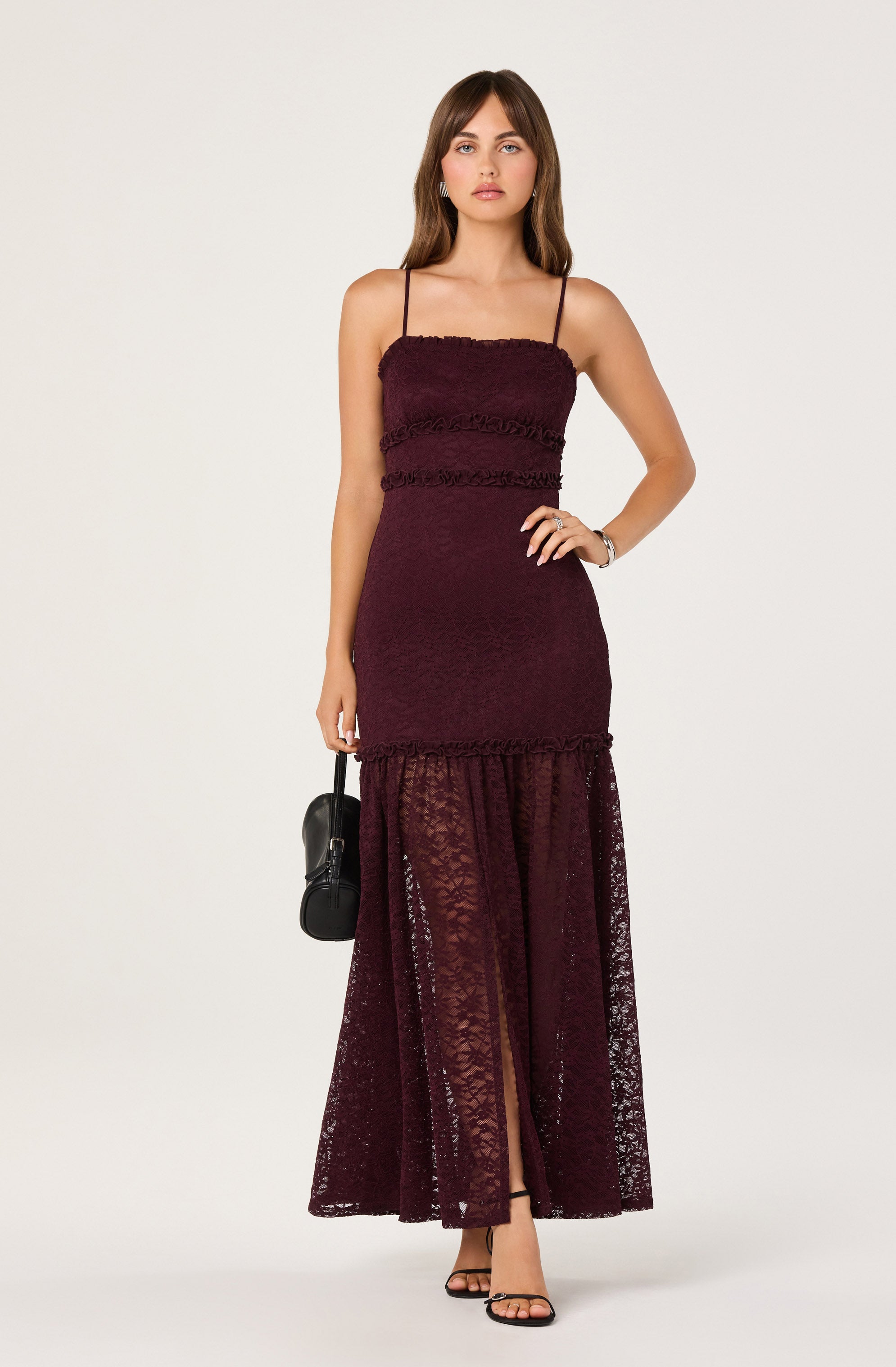 The model is wearing a sleeveless, burgundy dress with thin spaghetti straps. The dress has a fitted bodice with textured horizontal stripes and a sheer, lace overlay from the mid-thigh down to the floor, showing a slit at the front. The lace overlay features floral patterns and extends to the ground.