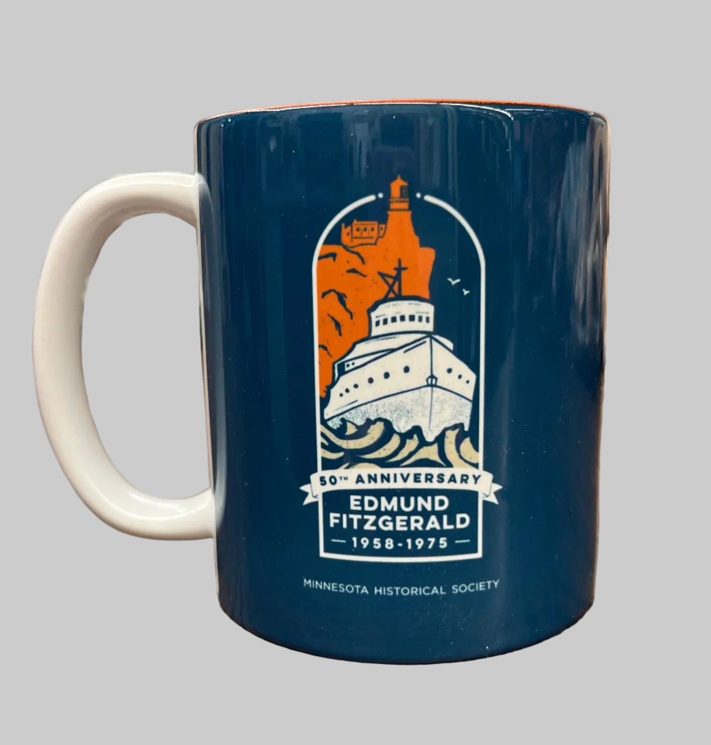The 50th Anniversary Edmund Fitzgerald Commemorative Mug is blue with a white handle, showing the ship on Lake Superior with waves and a lighthouse. It features “50th Anniversary Edmund Fitzgerald 1958-1975” and “Minnesota Historical Society.”.