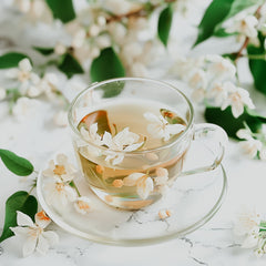 A cup of jasmine tea with visible jasmine flowers in the tea, placed on a saucer with a floral pattern, surrounded by more jasmine flowers and green leaves on a white background.
