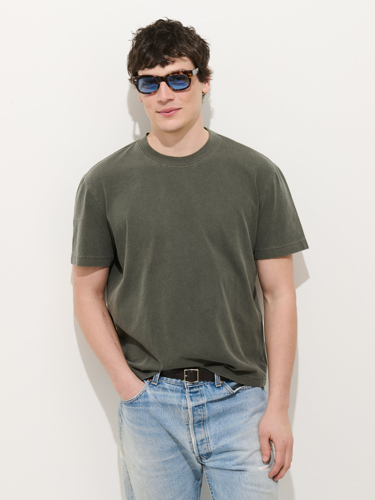 A young man wears blue sunglasses, light blue jeans, and the Crosby Tee in Midweight Jersey—an olive green, 100% cotton garment-dyed shirt—standing against a white background with one hand in his pocket.