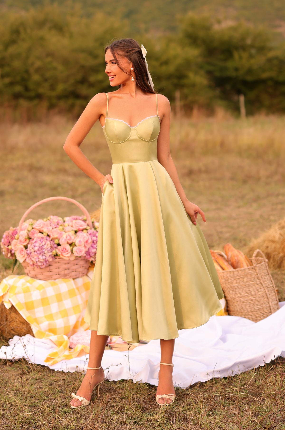 A woman in the Olivetta Olive Green Satin Midi Wedding Guest Dress stands outdoors on a picnic setup with flowers, bread, and a yellow-checkered cloth on the grass.