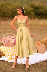 A woman in the Olivetta Olive Green Satin Midi Wedding Guest Dress stands outdoors on a picnic setup with flowers, bread, and a yellow-checkered cloth on the grass.