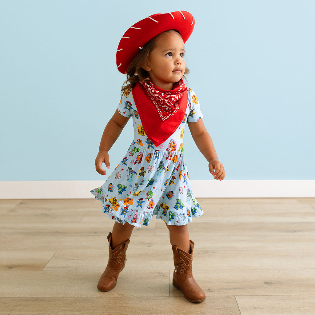 Child wearing a colorful dress with character patterns, a red bandana, and cowboy boots against a light blue wall.