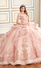 A woman stands indoors wearing the Quinceanera Dress PR30171 By Princesa, an elaborate off-the-shoulder pink gown with floral embroidery, glitter tulle, a tiara, and jewelry.