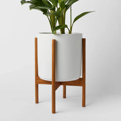 A white ceramic pot on a light wooden stand with four legs. Green plant is sitting inside the pot.