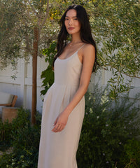 A woman with long dark hair stands outside by a white fence and green foliage, wearing the sleeveless light beige Rove Dress, made from fine Italian fabric, gazing calmly at the camera.